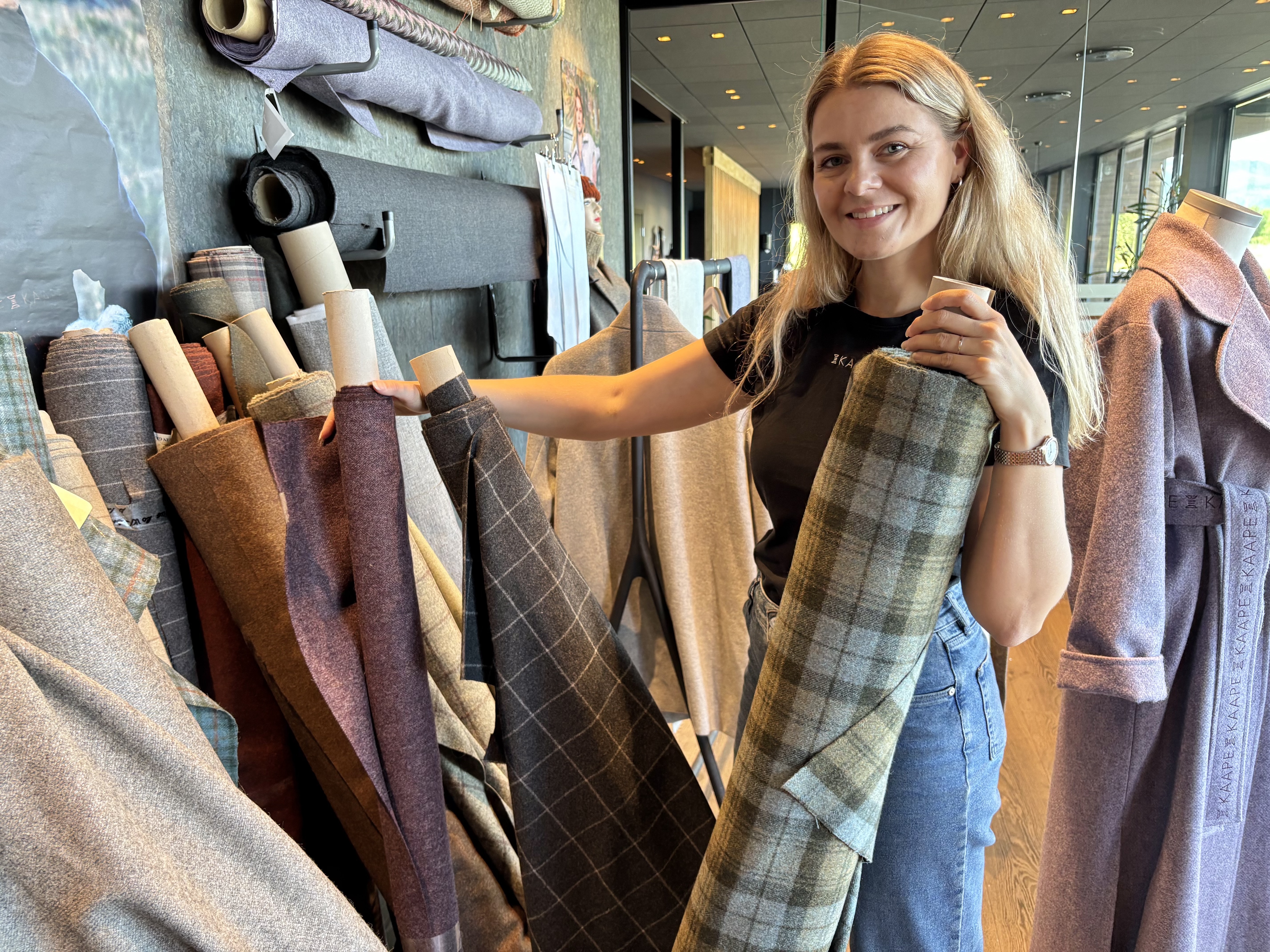 – Ei kåpe skal vera både varm og luftig og praktisk og slitesterk – og så fin at du ikkje vil ta ho av deg, meiner Kornelia. Her står ho med noko av det store stoffutvalet som er henta frå møbelindustrien i distriktet. Foto: Heidi Molstad Andresen / NPK